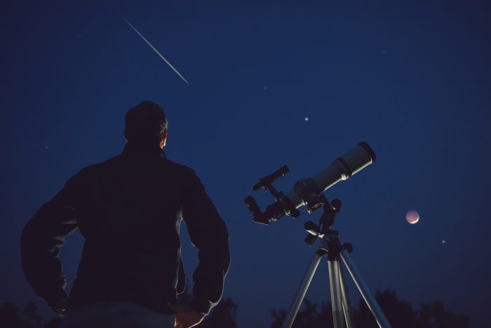 Silhouette of a man, telescope, stars, planets and shooting star under the night sky.